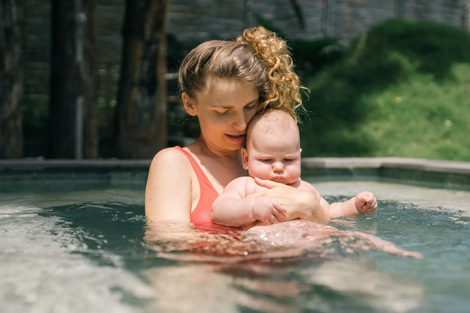 A loving mother holds her baby in a swimming pool, sharing a tender outdoors moment.